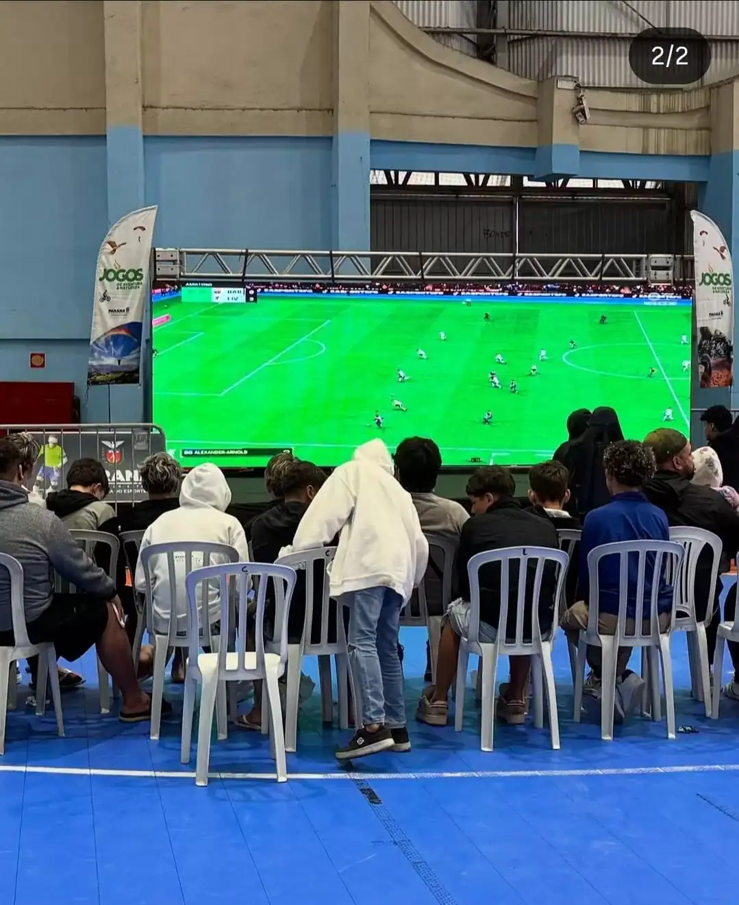 Tela de televisão exibindo jogo de futebol digital em evento esportivo, com público assistindo sentado em cadeiras na frente, promovendo entretenimento e tecnologia em esportes digitais.